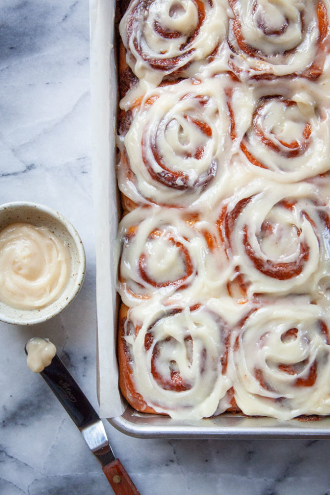A pan of cinnamon rolls frosted with cream cheese frosting, with a small bowl of extra frosting on the side as well as a small offset spatula with a dollop of frosting on it.