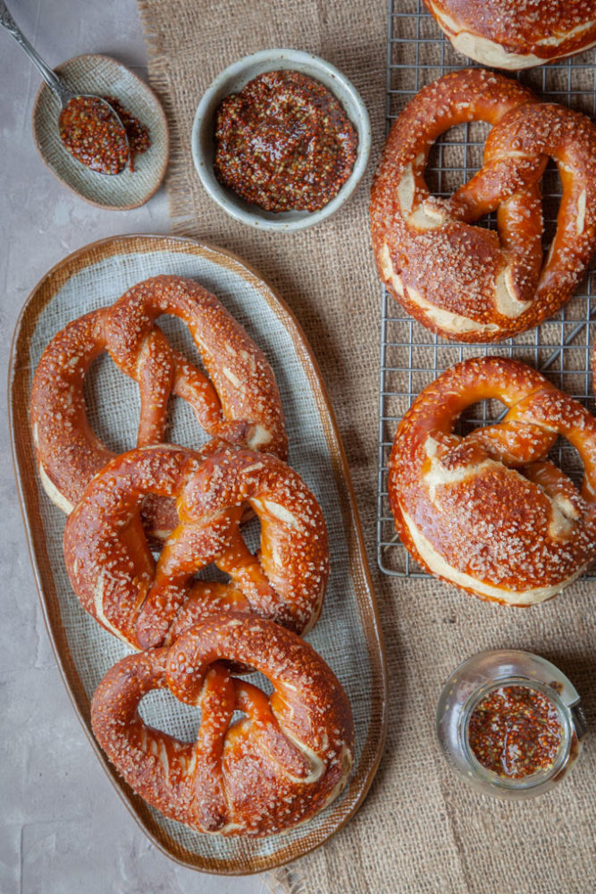 German-style pretzels on a plate with more pretzels on a wire cooling rack next to them. Bowls of mustard to dip into are next to the pretzels.