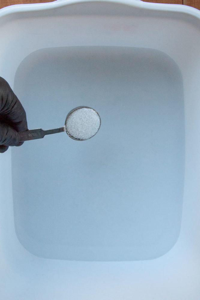 a hand with a tablespoon of lye powder over a plastic container of water.