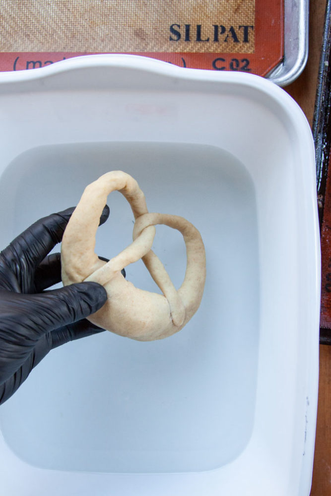 A hand wearing a glove, holding a pretzel about to dip it into a lye solution.