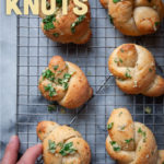 Fresh baked garlic knots sitting on a wire rack with a hand reaching out and grabbing one of them.