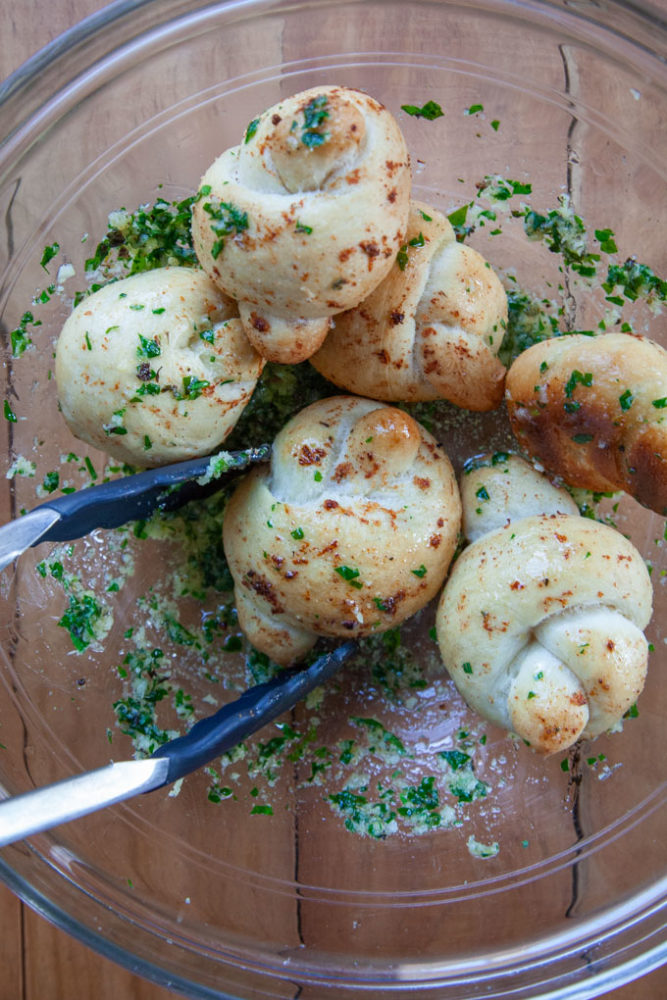 Garlic knots being tossed in parsley garlic butter in a bowl with tongs.