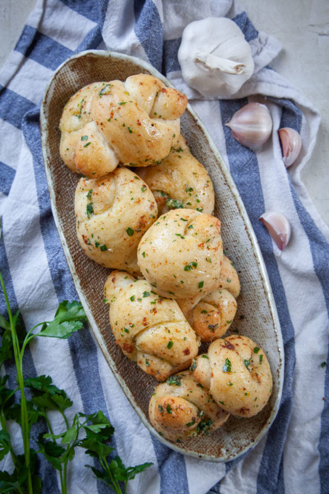 A pile of garlic knots in a plate, sitting on a blue striped kitchen towel, with a head of garlic next to it and Italian parsley next to it as well.