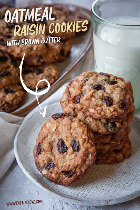 Oatmeal raisin cookies stacked on a plate, with a pile of cookies and a glass of milk in the background.