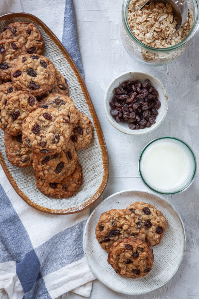 A pile of oatmeal raisin cookies on two plates with a glass of milk, a bowl of raisins and a container of rolled oats next to them.