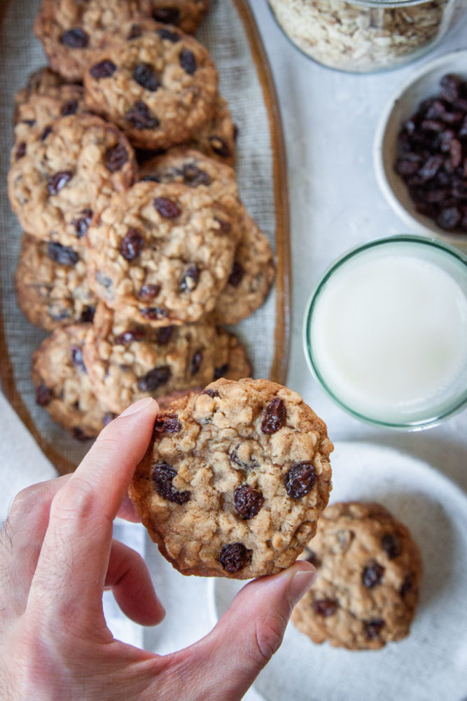 A hand lifting up an oatmeal raisin cookie with plates of cookies underneath it.