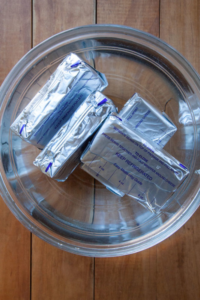 Four bricks of cream cheese in their aluminum foil wrapper sitting in a bowl of warm water.