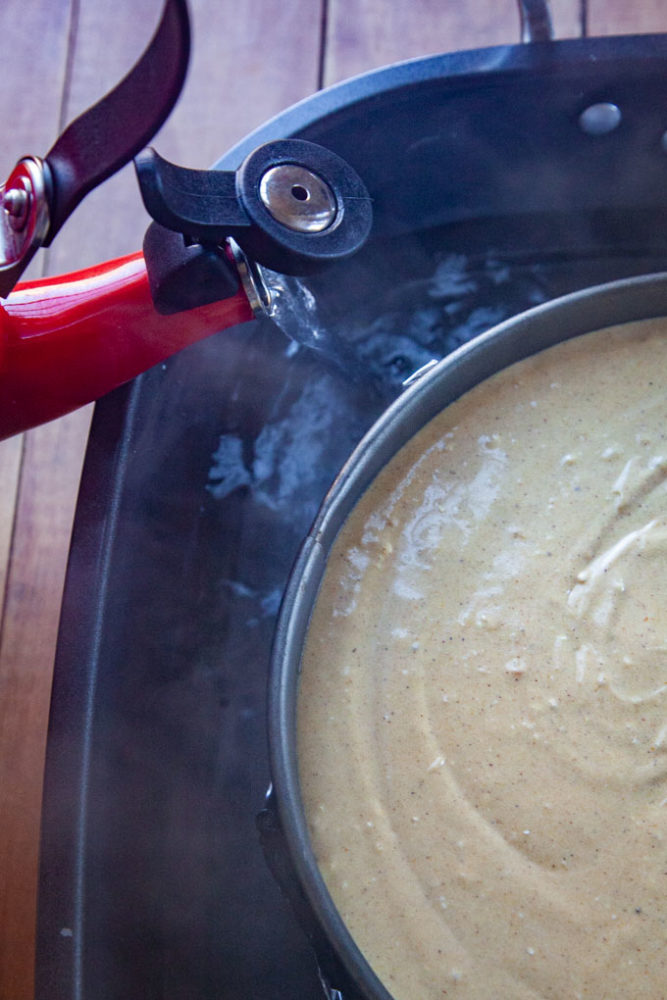 A water kettle pouring boiling water into a roasting pan that has a springform pan with an unbaked pumpkin cheesecake in it.