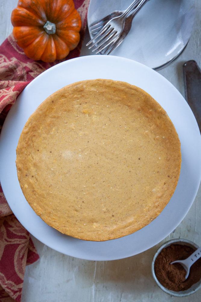 A whole pumpkin cheesecake on a serving plate with a small pumpkin and a bowl of pumpkin pie spice blend next to it.