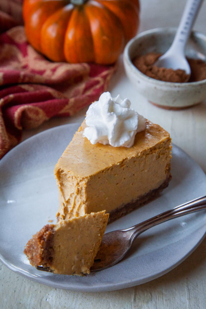 A slice of pumpkin cheesecake with whipped cream on it and a bowl of pumpkin pie spice and a pumpkin in the background.