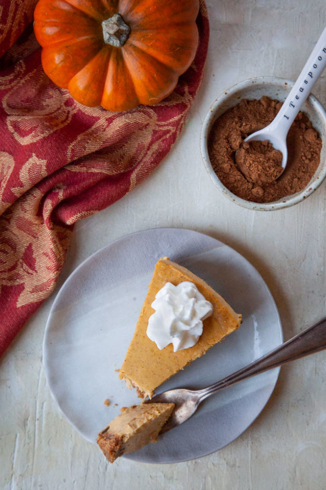 A slice of pumpkin cheesecake with whipped cream on it and the remaining cheesecake, a small pumpkin and a bowl of pumpkin pie spice next to it.