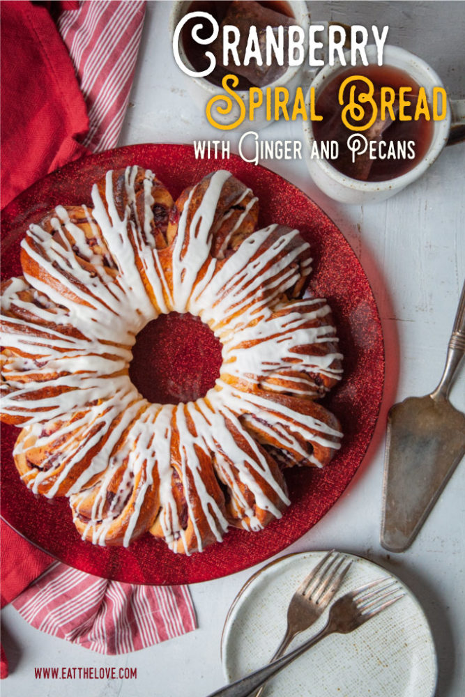 A large cranberry spiral bread on a red plate with two small desserts plates and two mugs of hot tea next to it.