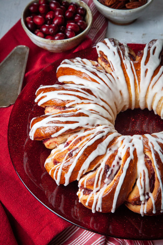 A large cranberry spiral bread on a red plate with a small bowl of cranberries and pecans behind the plate.