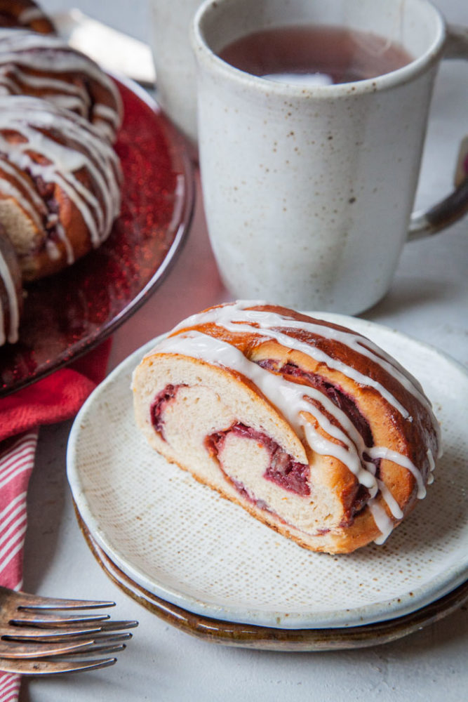 A slice of spiral cranberry bread on a plate with the remaining bread behind it on a red plate and a mug of tea behind the slice.