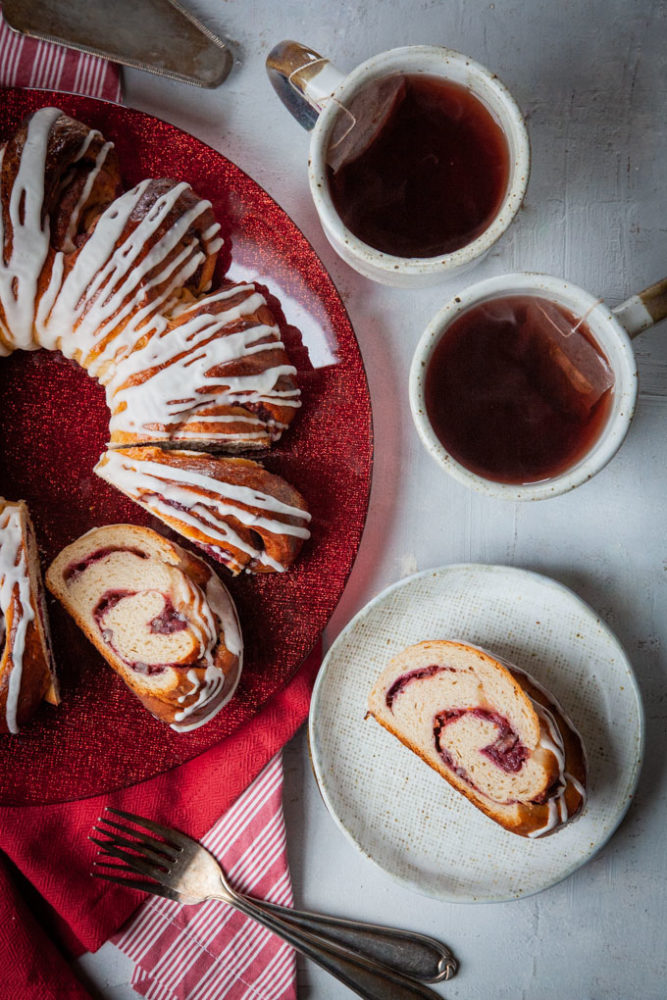 A slice of cranberry swirl bread on a plate next to the remaining cranberry bread and two mugs of tea next to it.