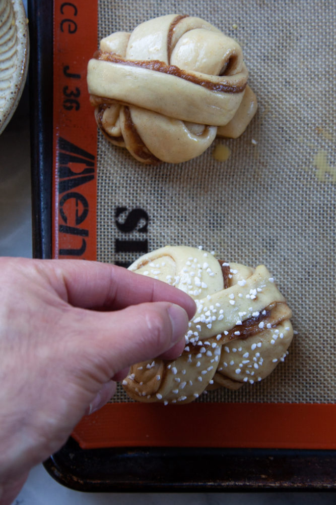 A hand sprinkling Swedish pearl sugar over an unbaked cardamom bun on a baking sheet.