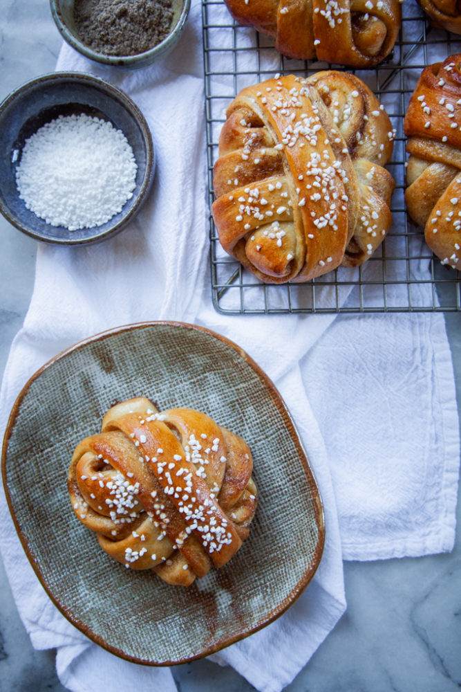 A cardamom bun on a plate, with two small plates next to it filled with Belgian pearl sugar and cardamom. A wire cooling rack with more buns is next to the small plates.