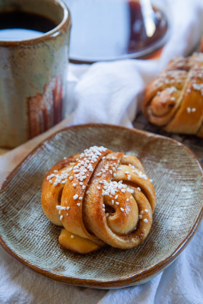 A cardamom on a plate with a mug of coffee and a Chemex coffee maker behind it, along with a wire cooling rack with more buns on them.