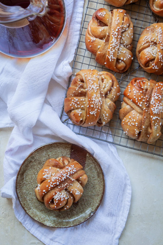 A cardamom on a plate with a Chemex coffee maker behind it and a wire cooling rack with more buns on them.