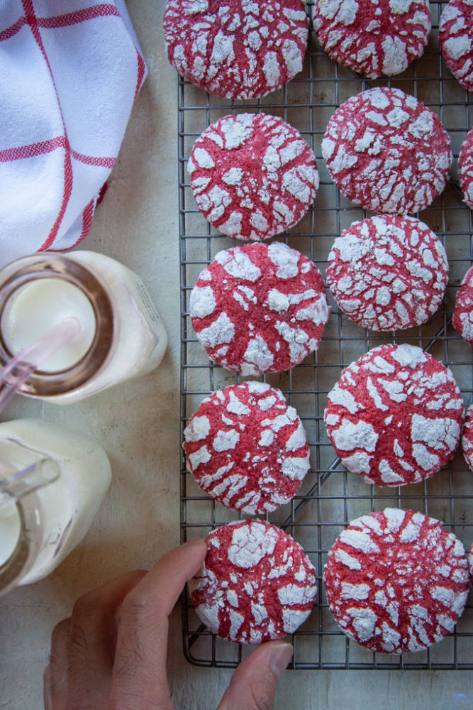A hand reaching for a red velvet crackle cookie on a wire cooling rack. Next to the hand are two milk bottles with milk in them.