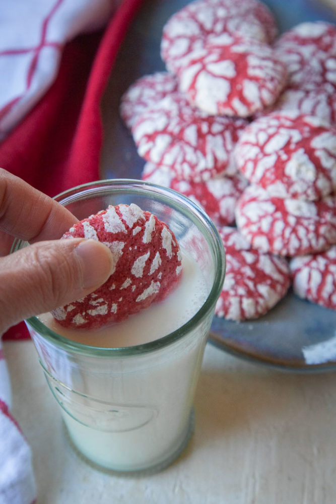 A hand dunking a red velvet crackle cookie into a glass of milk with a plate of cookies behind it.
