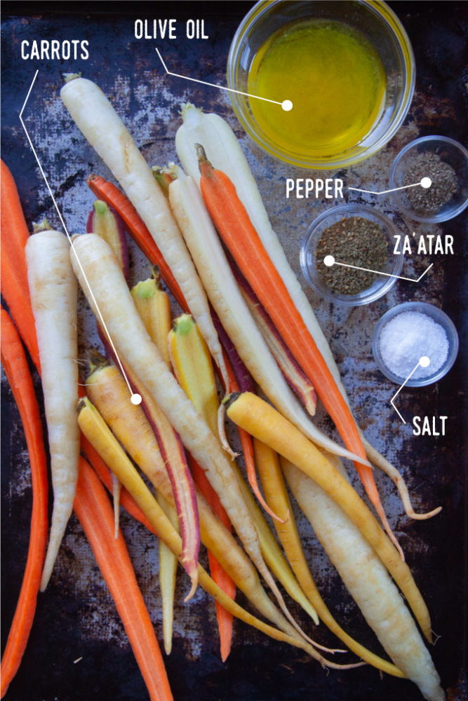 Rainbow carrots sliced lengthwise on a rimmed baking sheet with small bowls filled with olive oil, salt, pepper and za'atar spices.