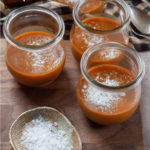 Jars of salted caramel pots de creme on a cutting board with a small dish of flaky sea salt next to them.