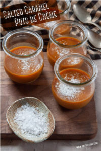 Jars of salted caramel pots de creme on a cutting board with a small dish of flaky sea salt next to them.