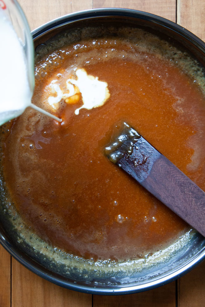 Hot cream being poured into a pan filled with caramelized sugar and a wooden spatula in it.