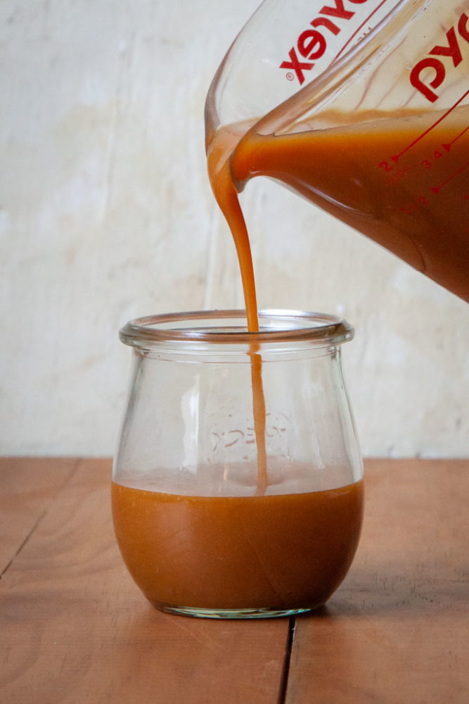 a glass measuring cup pouring caramel custard into a small glass jar.