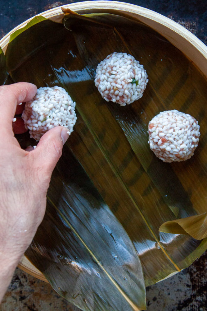 A hand placing an uncooked Chinese pearl meatball in bamboo steamer basket lined with bamboo leaves.
