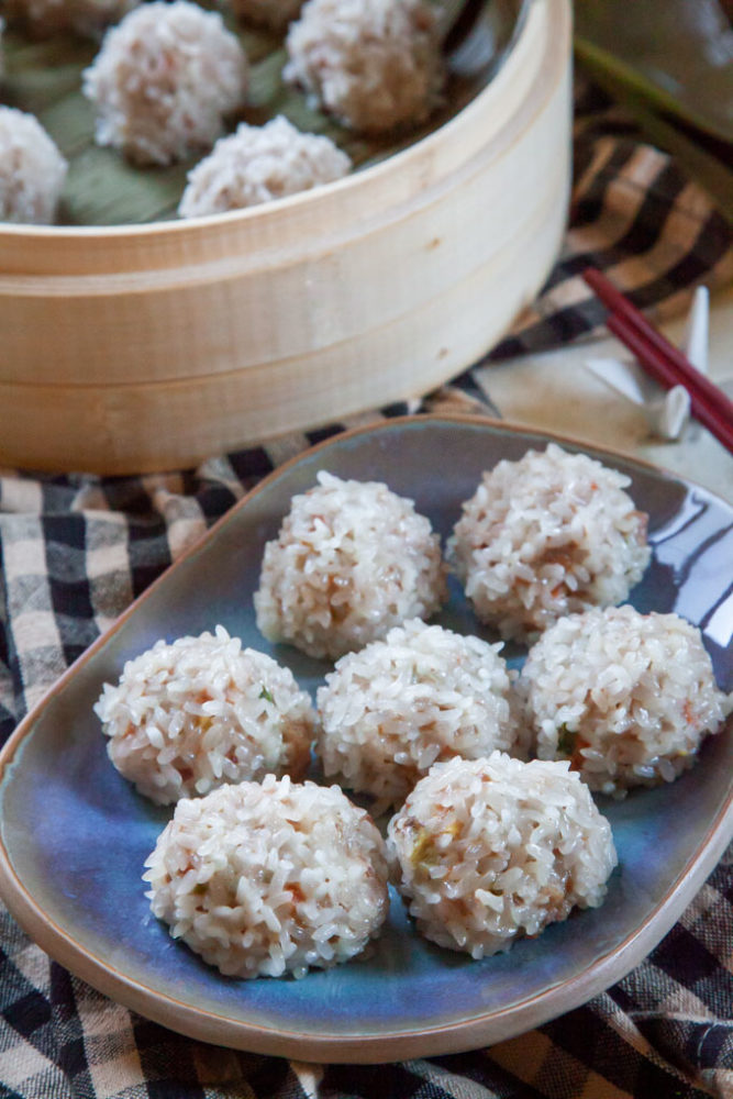 Chinese pearl meatballs on a blue plate sitting on a cloth checked napkin with a bamboo steamer basket of more pearl meatballs.