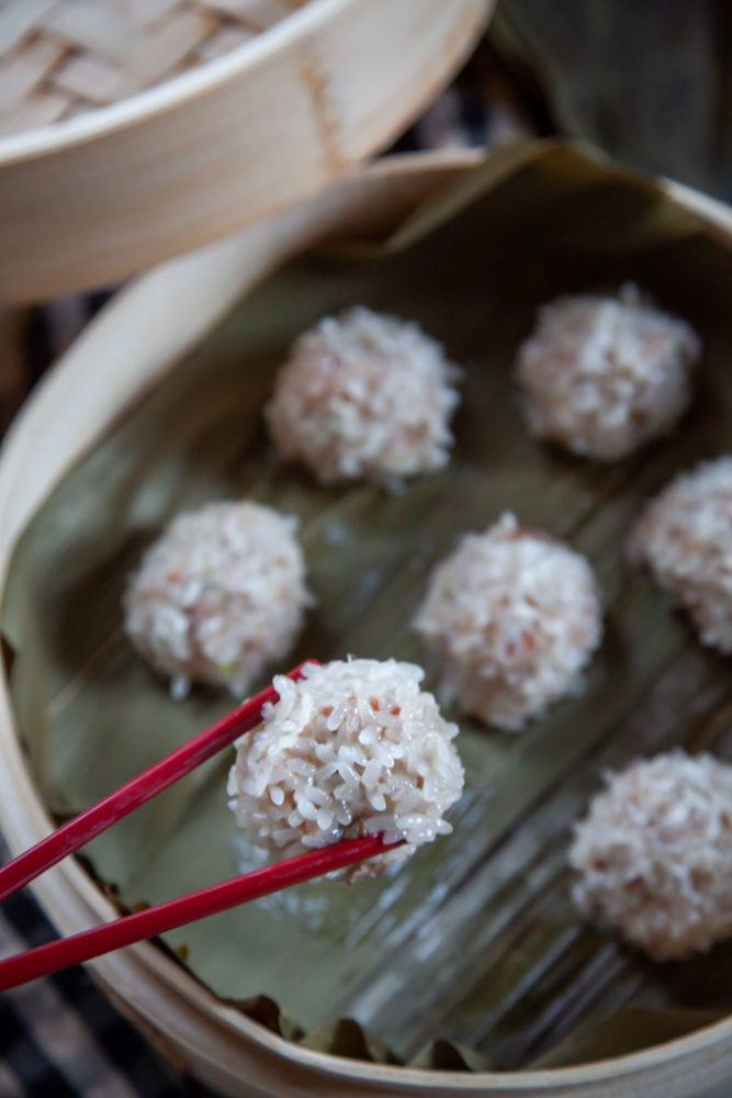 Red chopsticks holding up a Chinese pearl meatball, with a bamboo steamer basket of pearl meatballs underneath it.