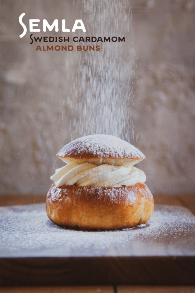 Semla, a Swedish cardamom almond bun on a cutting board with a dusting of powdered sugar being sprinkled on it.