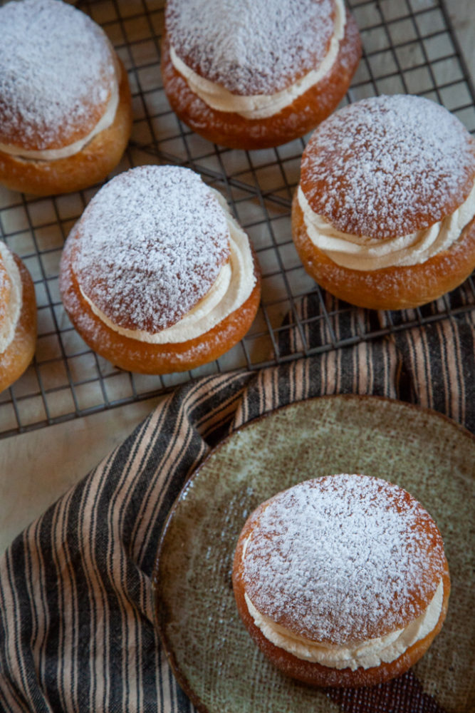 A Swedish semla bun sitting on a plate with more semla buns behind it on a wire rack.