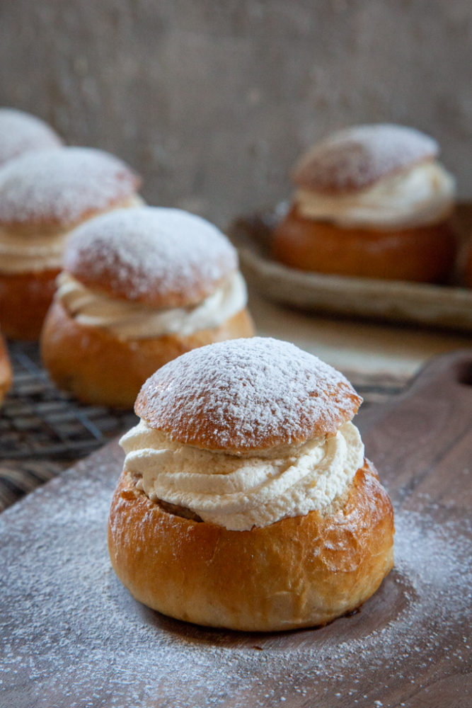 A Swedish semla bun on a cutting board dusted with powdered sugar. More semlor buns are in the background sitting on a wire rack and another plate.