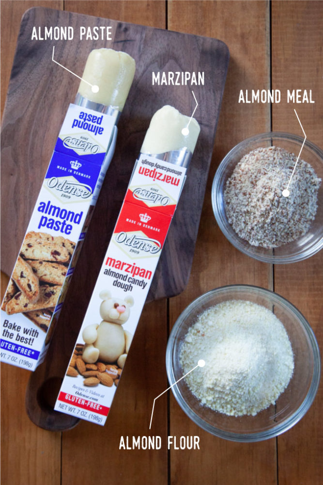 A tube of almond paste and marzipan on a wooden cutting board, with a bowl of almond meal and a bowl of almond flour next to the cutting board.