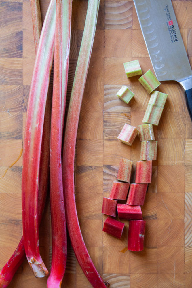 Stalks of rhubarb on a cutting board, with one rhubarb stalk cut into pieces with a knife next to the pieces.