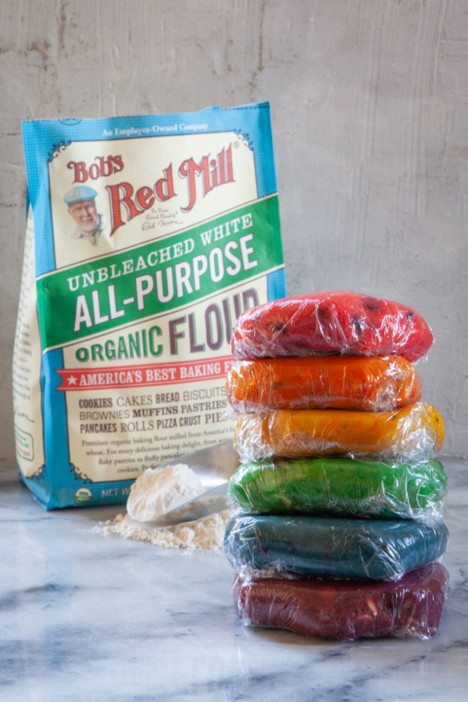 A stack of colored cookie dough, wrapped in plastic with a bag of Bob's Red Mill behind it and a scoop of flour on the table.