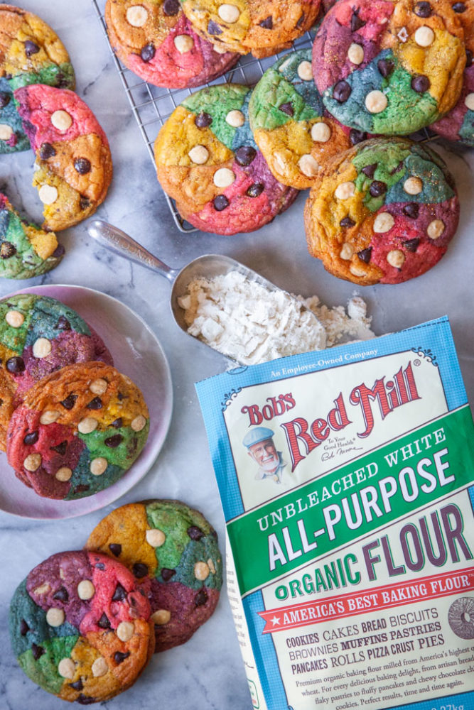 Rainbow chocolate chip cookies with a bag of Bob's Red Mill all-purpose flour next to them and a scoop of flour on the table.