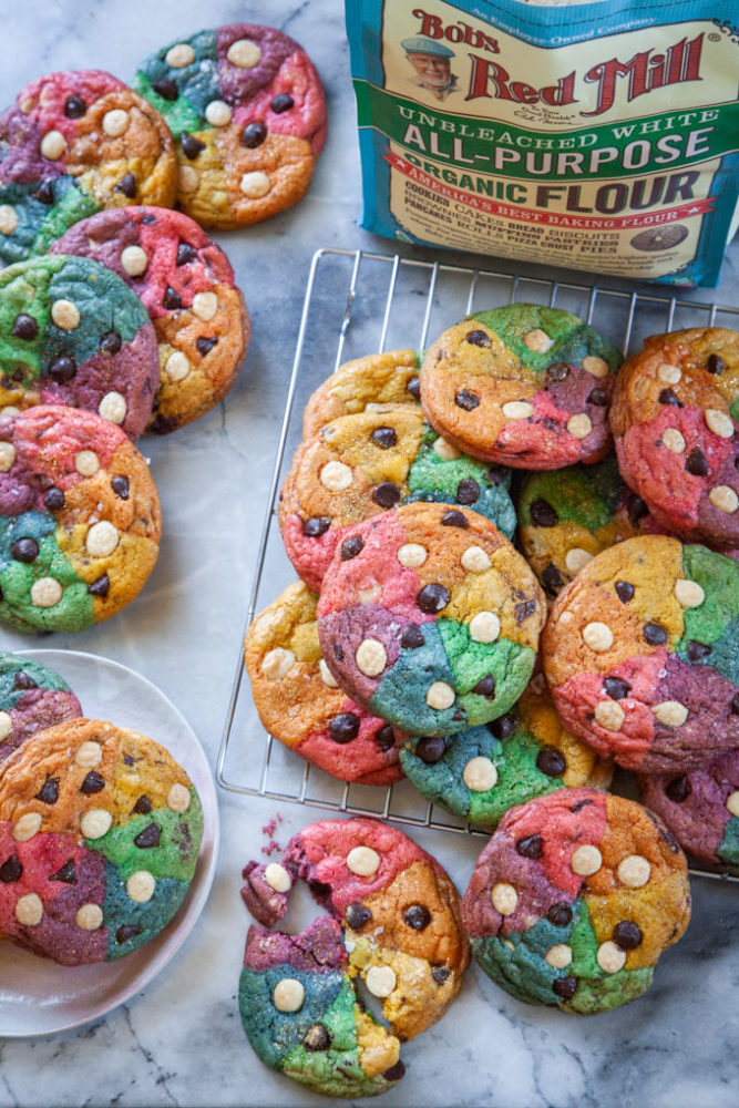 Rainbow chocolate chip cookies on a wire rack and on a plate, next to a bag of Bob's Red Mill all-purpose flour.