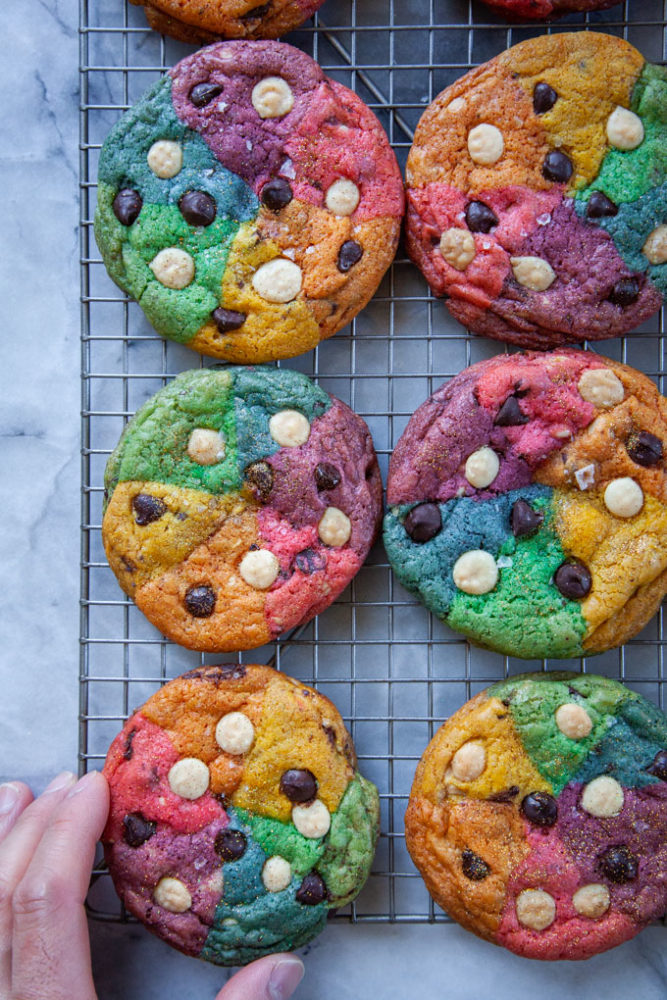 A hand reaching for a rainbow chocolate chip cookie cooling on a wire rack.