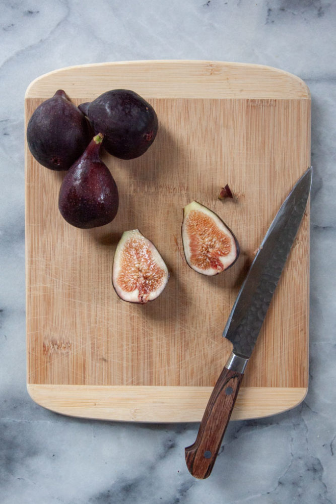 Fresh figs on a cutting board with one fig cut in half. A knife is next to the cut fig.
