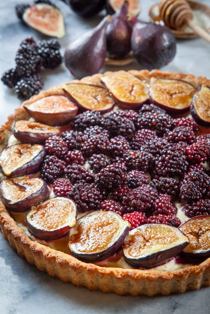 A fig and blackberry tart on a marble surface, with fresh figs, blackberries and a honey dipper with some honey next to it.