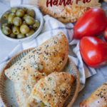 Olive, tomato and onion hand pies on a plate, surrounded by tomatoes and a bowl of more olives.