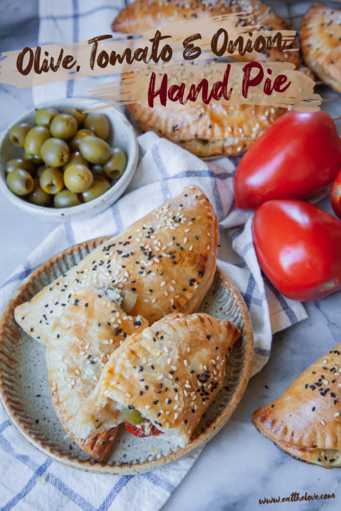 Olive, tomato and onion hand pies on a plate, surrounded by tomatoes and a bowl of more olives.