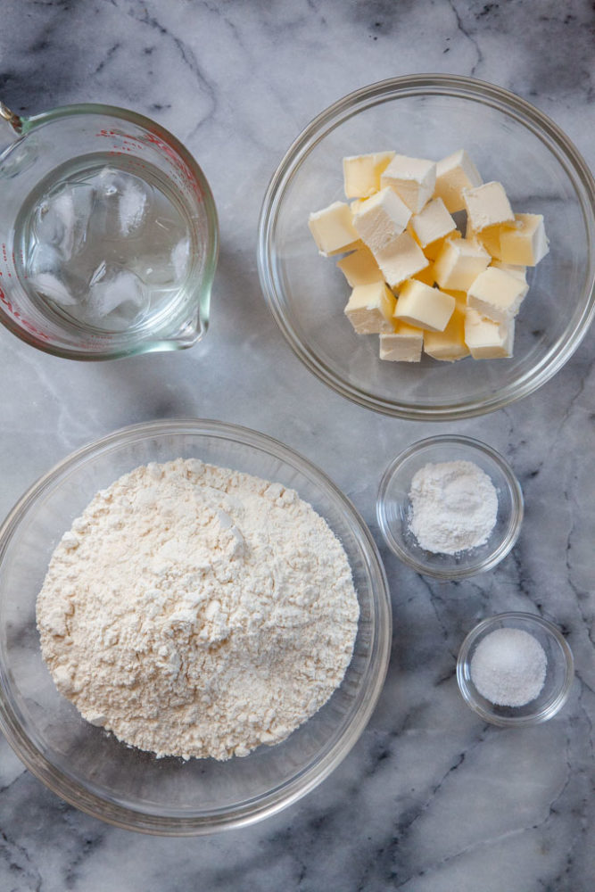 Ice water, cubed butter, flour, salt and baking powder in bowls on a marble surface.