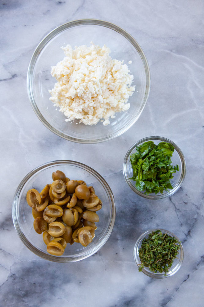 Feta, cut green olives, parsley and thyme in small bowls on a marble surface.