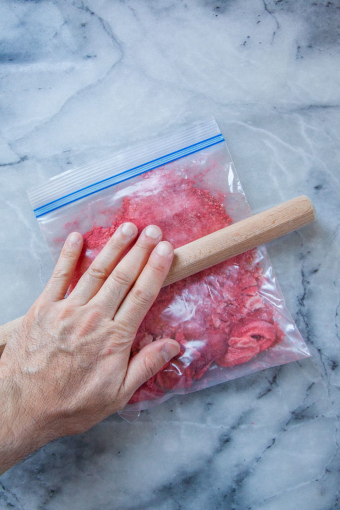 A hand crushing freeze dried strawberry slices in a Zipoc bag with a rolling pin.