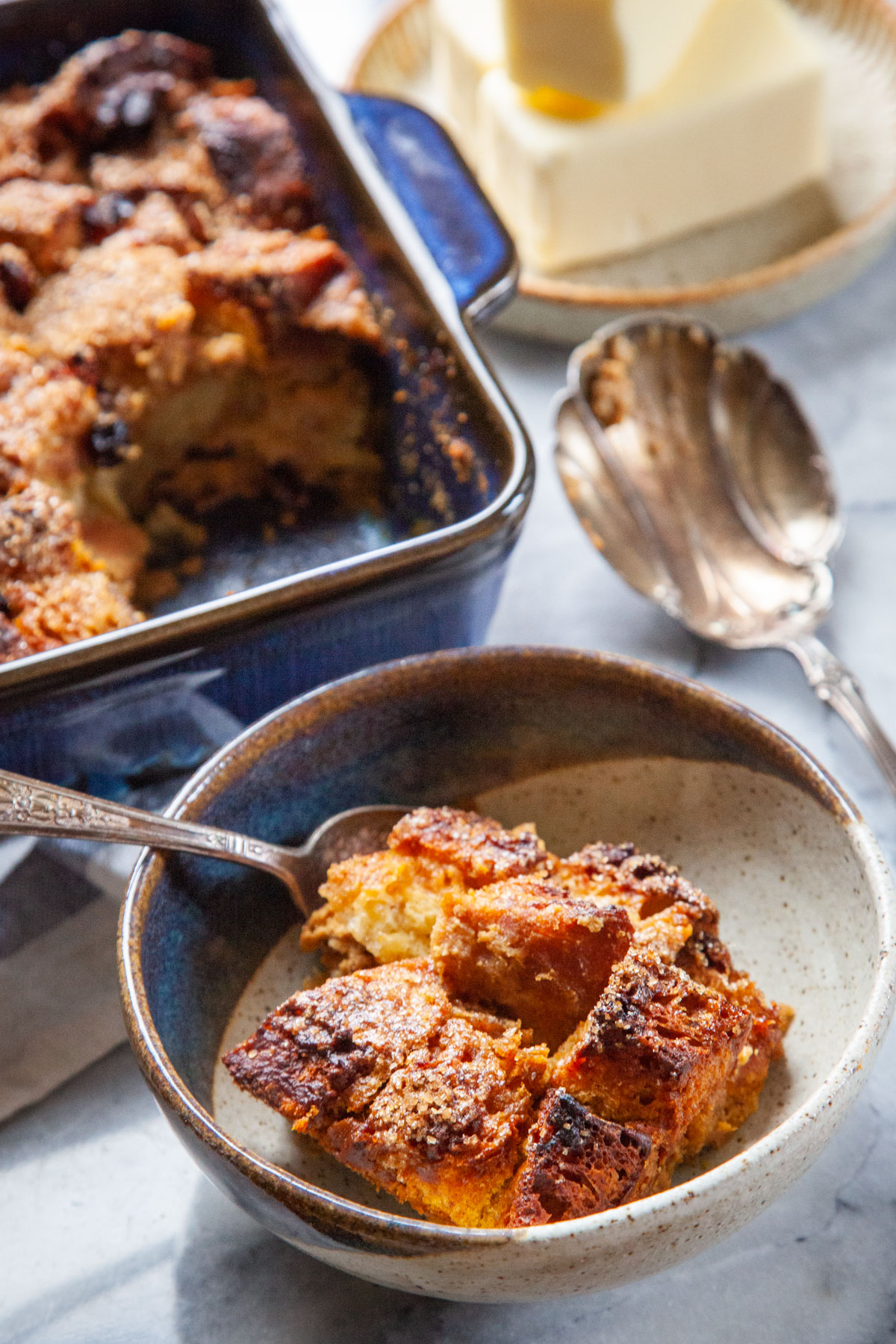 A bowl of pumpkin bread pudding with a pan of more bread pudding and a plate of butter sticks near it.